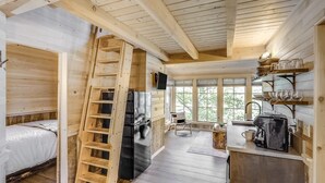 Interno di una baita con una scala in legno, un letto, angolo cottura con frigorifero e finestra con vista sul verde.