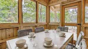 A wooden cabin with a dining table set for four, surrounded by large windows offering a view of a forest. - Outdoor Shower Treehouse in Smokies | Cottonwood (Sevierville)