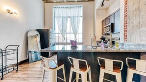 A kitchen with a central island, bar stools, a window with sheer curtains, and a brick accent wall. - Sosuite | 2BR Apt w W/D, Gym, On-Site Restaurant (Philadelphia)
