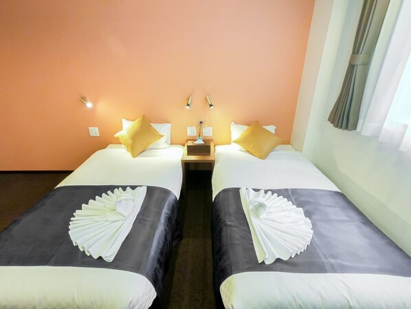 Two single beds with white pillows and black bedspreads, a nightstand with a lamp and table, and a window with sheer curtains.
