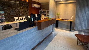 Una moderna zona de recepción con pared de piedra, mostrador de madera, máquina de café y área de descanso con silla de mimbre.