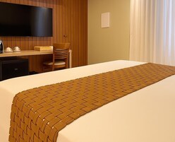 Una habitación de hotel con cama, escritorio, silla, televisor y ventana con cortinas.