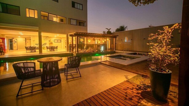 Un área de piscina moderna al aire libre con terraza de madera, sillas de descanso y un comedor con paredes de vidrio.