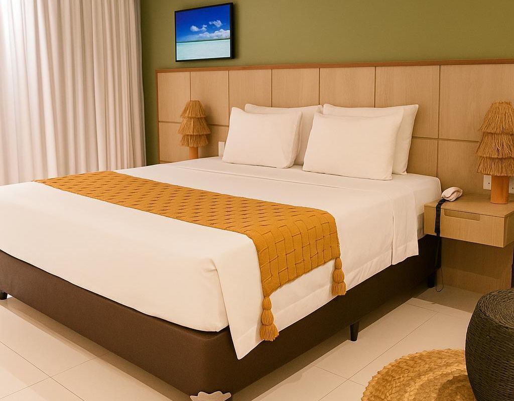 A hotel room with a large bed, a wooden headboard, a nightstand with a lamp, and a view of the sea through a window.