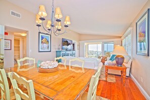 A spacious dining area with a wooden table and chairs, a chandelier, and a sofa with a lamp. - Tarpon Beach 112 offers the BEST Beach View! Stunning, Top Floor, Corner Unit (Sanibel)