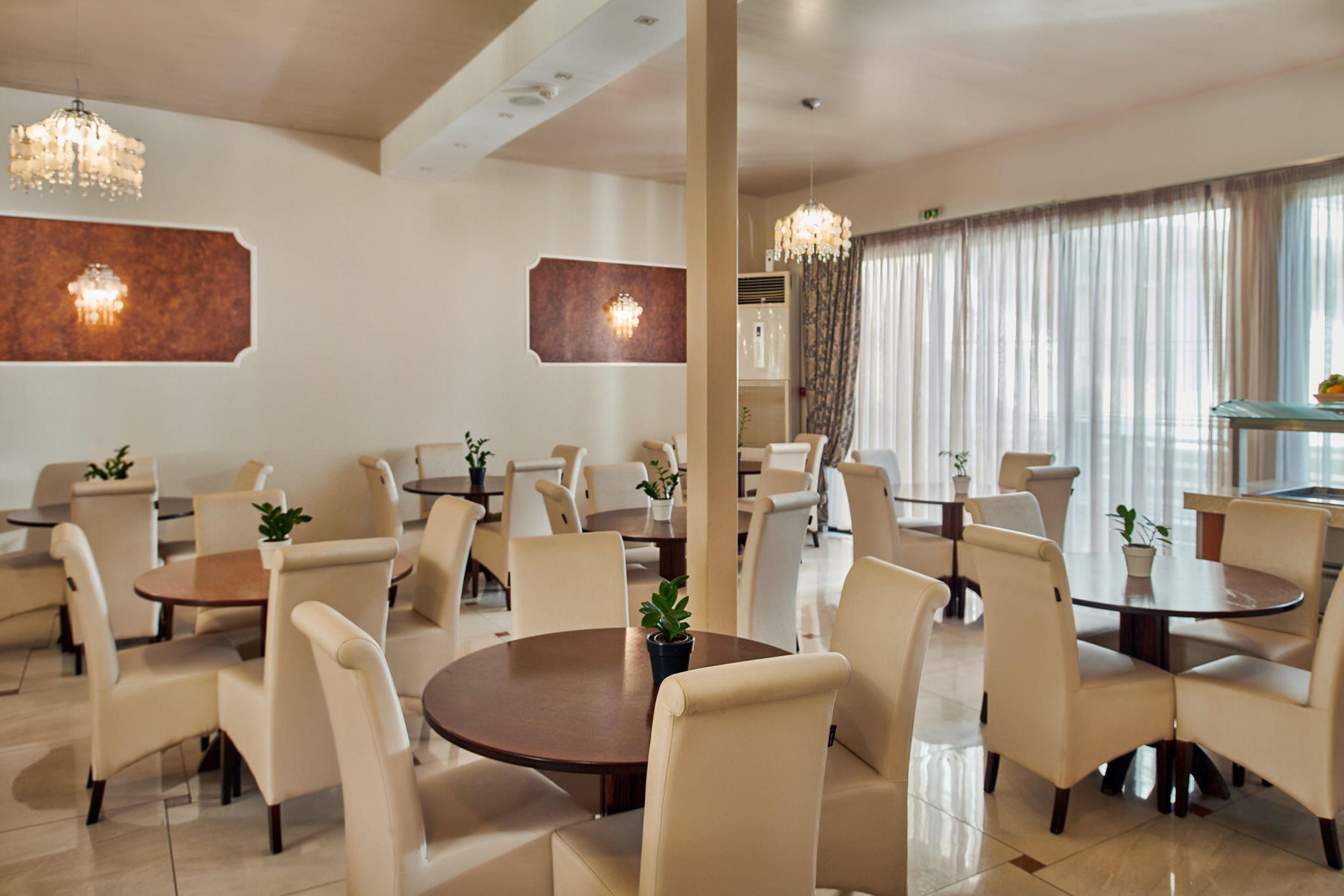 A hotel dining area with round tables, beige chairs, and potted plants.