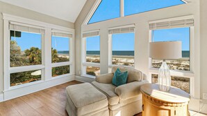 Ein Zimmer mit großem Fenster mit Blick auf einen Strand, ein beiges Sofa mit einem türkisfarbenen Kissen und ein Holztisch mit einer Lampe.