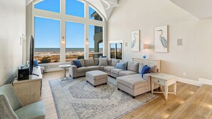 Ein geräumiges Wohnzimmer mit einem großen Bogenfenster und Blick auf den Strand, einem Ecksofa, einem kleinen Beistelltisch und einem dekorativen Teppich.