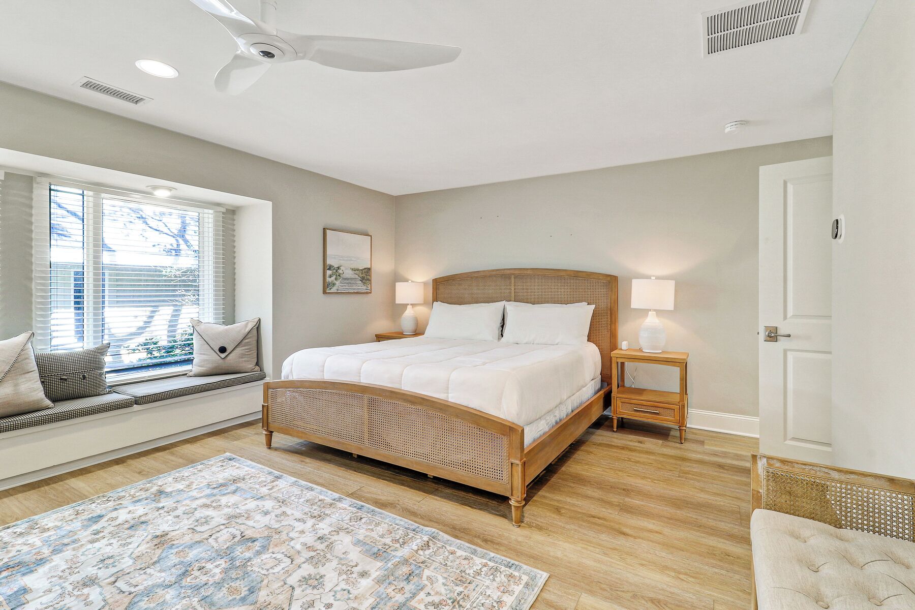 A bedroom with a large bed, bedside lamps, a window with blinds, a rattan headboard, and a patterned rug.