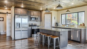 A modern kitchen with stainless steel appliances, wooden flooring, and a large island with bar stools. - Views for Days, New Cabin Near Terry Peak Ski Area w/ Hot Tub and Large Deck! (Lead)