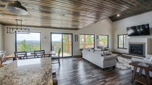A spacious living area with a large window, a fireplace, and a television mounted on the wall. - Views for Days, New Cabin Near Terry Peak Ski Area w/ Hot Tub and Large Deck! (Lead)