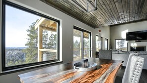 A modern dining area with a wooden table, large windows, and a view of the forest. - Views for Days, New Cabin Near Terry Peak Ski Area w/ Hot Tub and Large Deck! (Lead)
