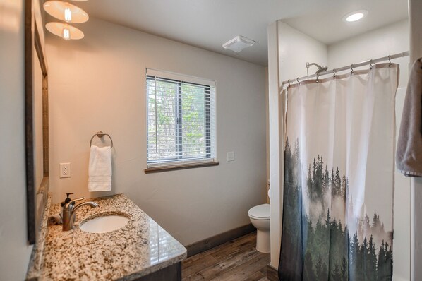 A bathroom with a granite sink, a window with blinds, a toilet, and a shower curtain with a forest design. - Views for Days, New Cabin Near Terry Peak Ski Area w/ Hot Tub and Large Deck! (Lead)