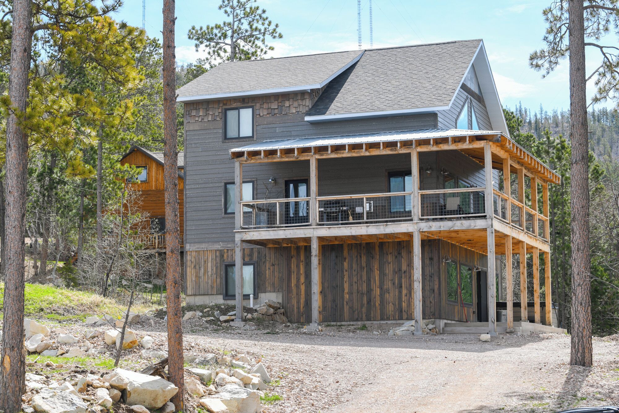 A multi-story wooden cabin with a balcony, surrounded by trees and a gravel driveway.