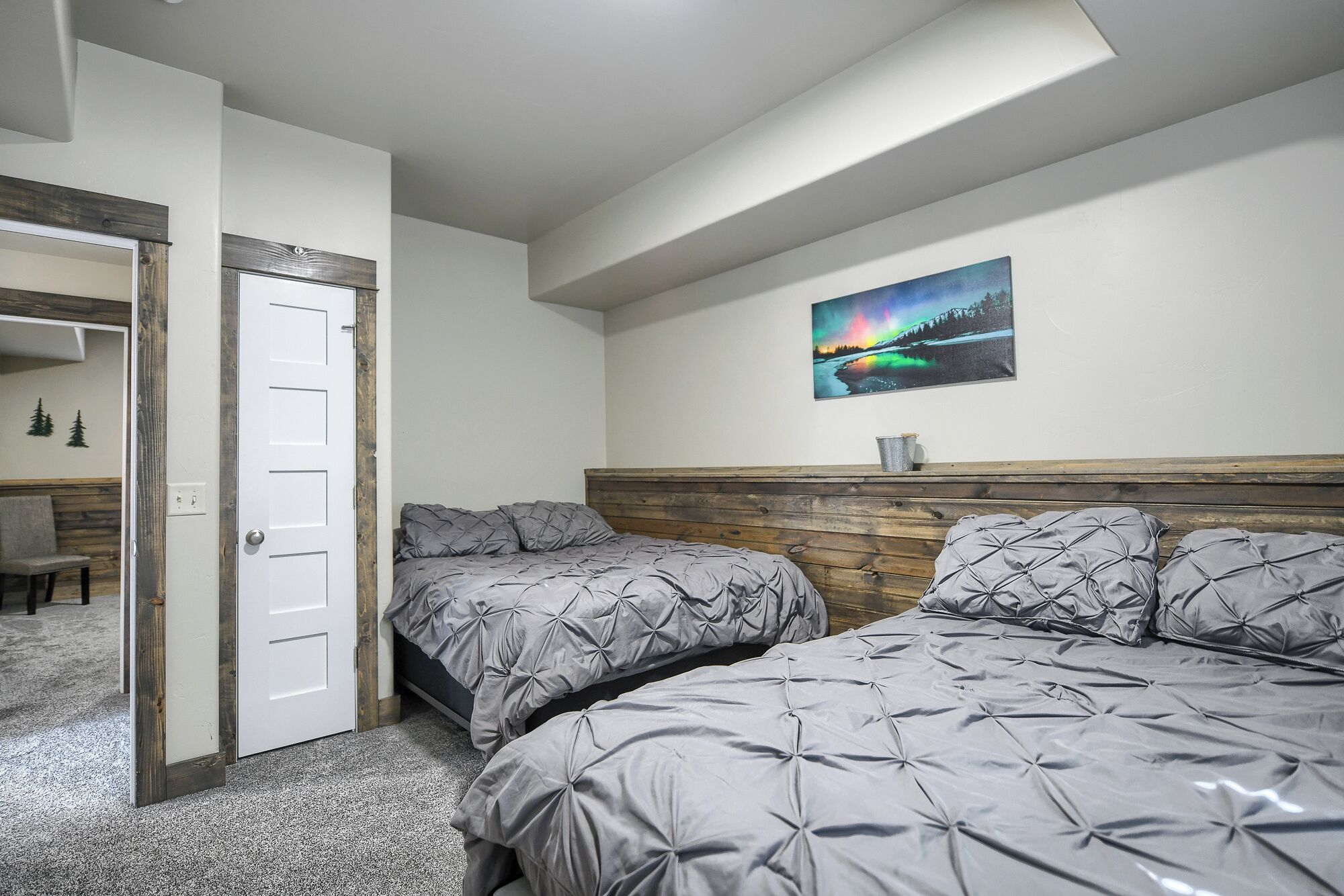 Une chambre avec deux lits, une tête de lit en bois, un tableau accroché au mur et une porte donnant sur une autre pièce.