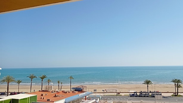 A beach view from a balcony overlooking a sandy beach with palm trees, a restaurant, and a clear blue sky. - Peñíscola Playa Inmomart (Peñiscola)