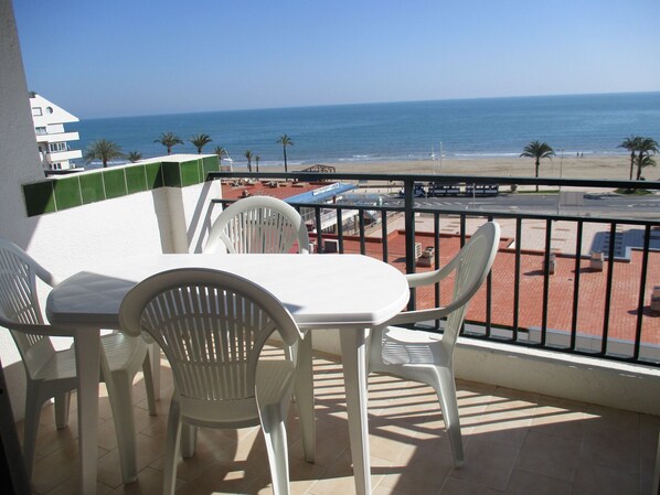 A balcony with a table and chairs, overlooking a beach and a parking area.