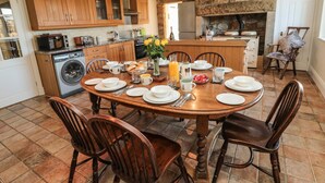 Un coin repas avec une table ronde en bois, dressée pour six personnes, entourée de chaises en bois. Au fond de la pièce, on aperçoit une cheminée en pierre, et la pièce est équipée de meubles en bois et d’un lave-linge.