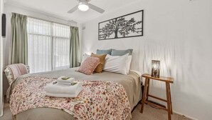 Une chambre à coucher avec un lit, un ventilateur de plafond, une table de chevet avec une lampe et un tableau représentant un arbre accroché au mur.