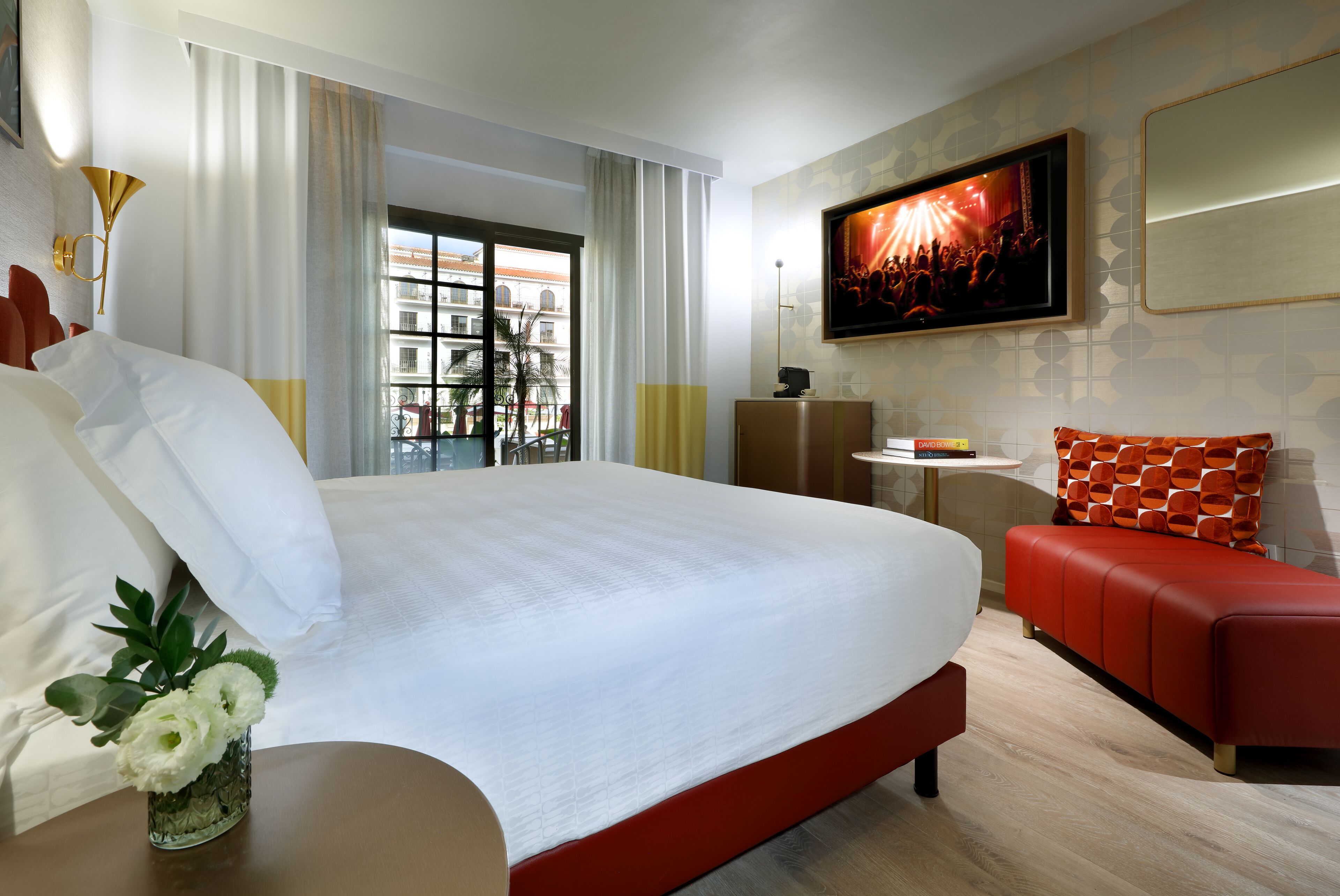 Une chambre d’hôtel avec un grand lit, un canapé rouge, un téléviseur fixé au mur et une fenêtre avec des rideaux.