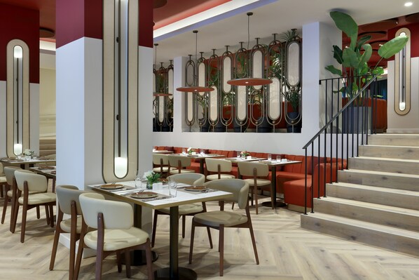 A modern restaurant with a herringbone wood floor, white tables, and beige chairs. There are multiple dining tables arranged in the center, surrounded by red cushioned seating. A staircase with a black railing leads to an upper level.