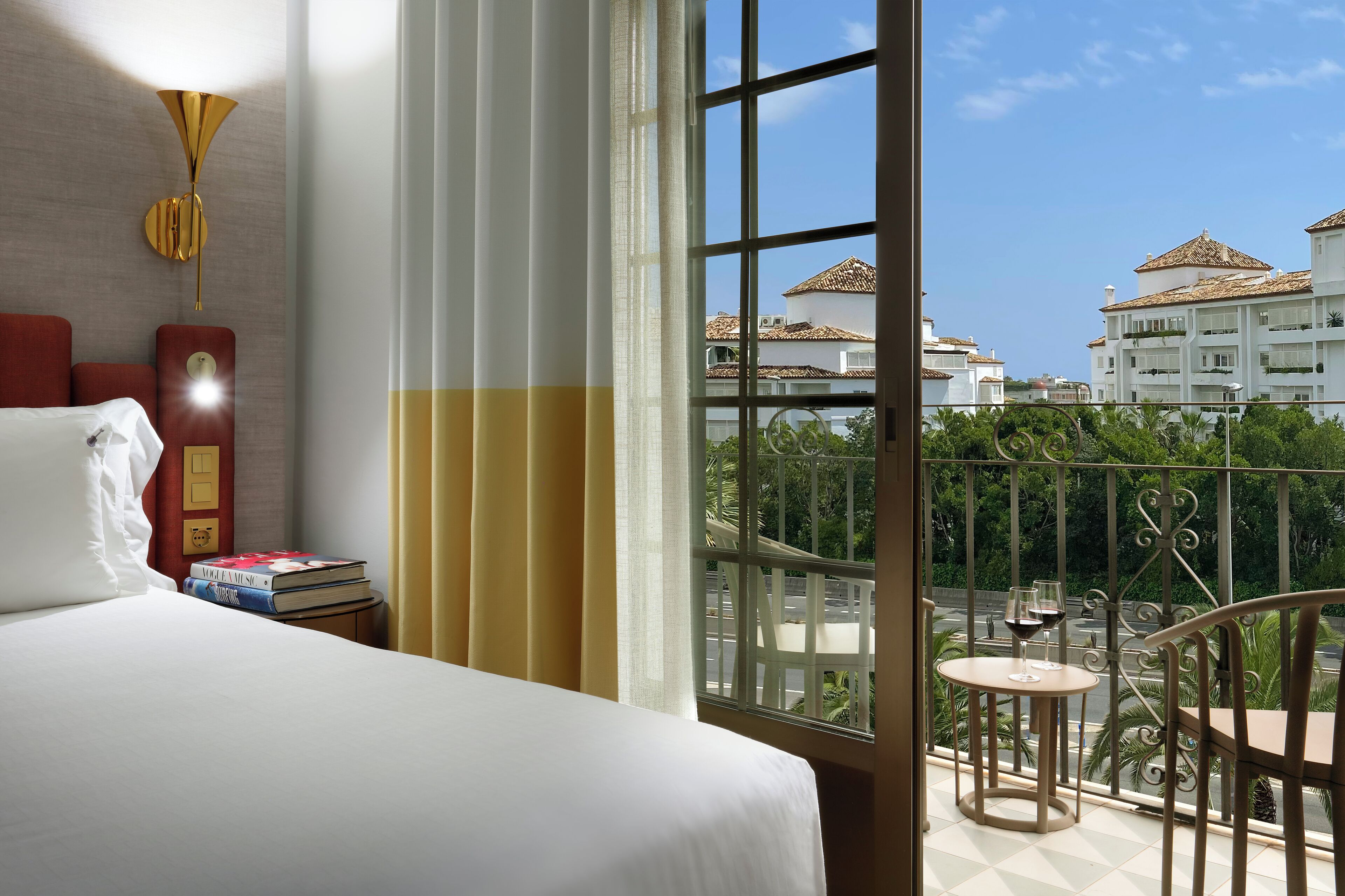 A hotel room with a bed, a bedside table with books, a window with a view of buildings and greenery, and a balcony with a table and chairs.