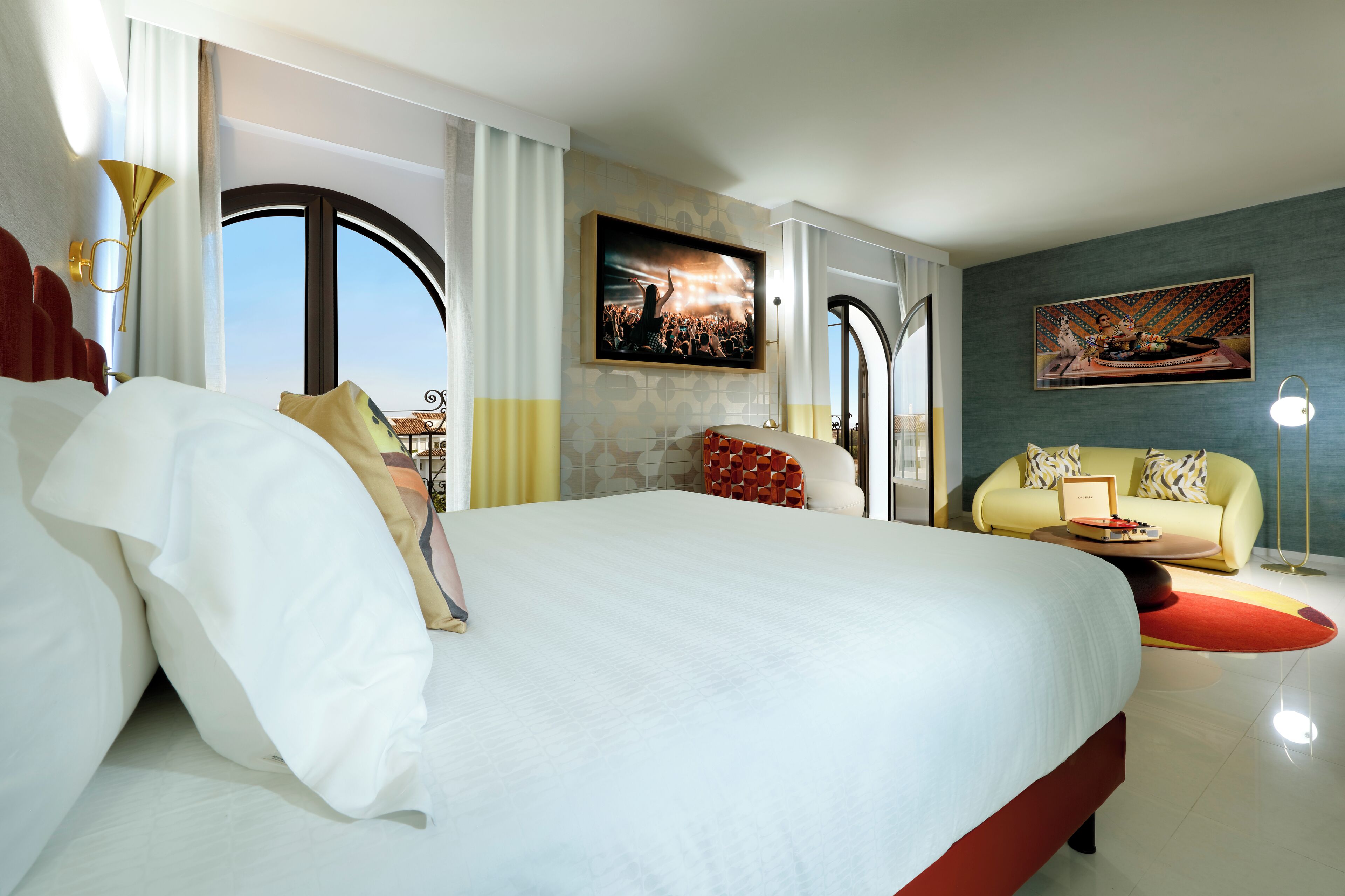 Habitación de hotel con una cama grande, un sofá y una mesa de comedor. Hay dos cuadros en las paredes y una ventana con cortinas.