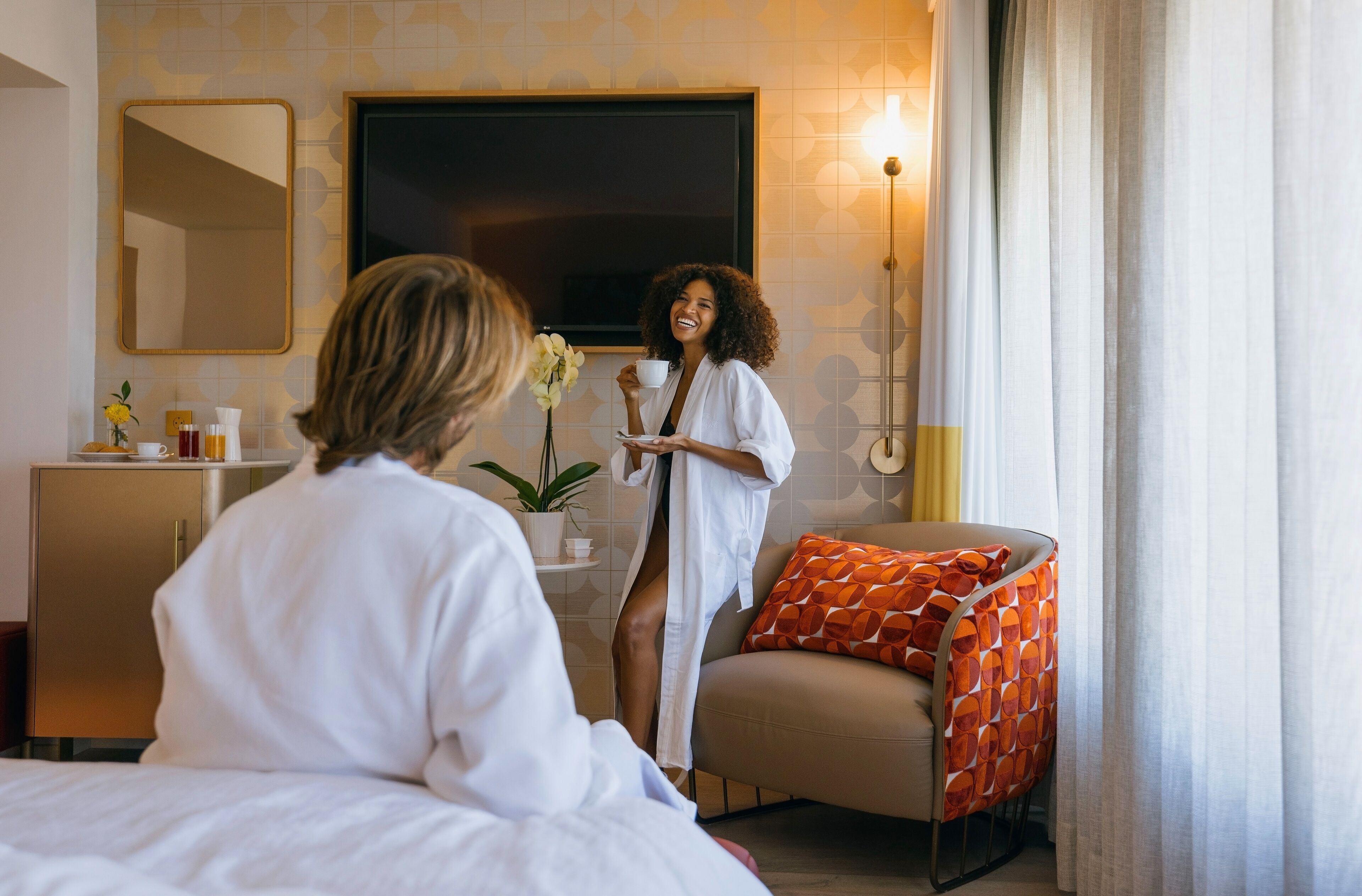 A hotel room with a bed, a bedside table with toiletries, a mirror, a TV, and a chair with an orange cushion.