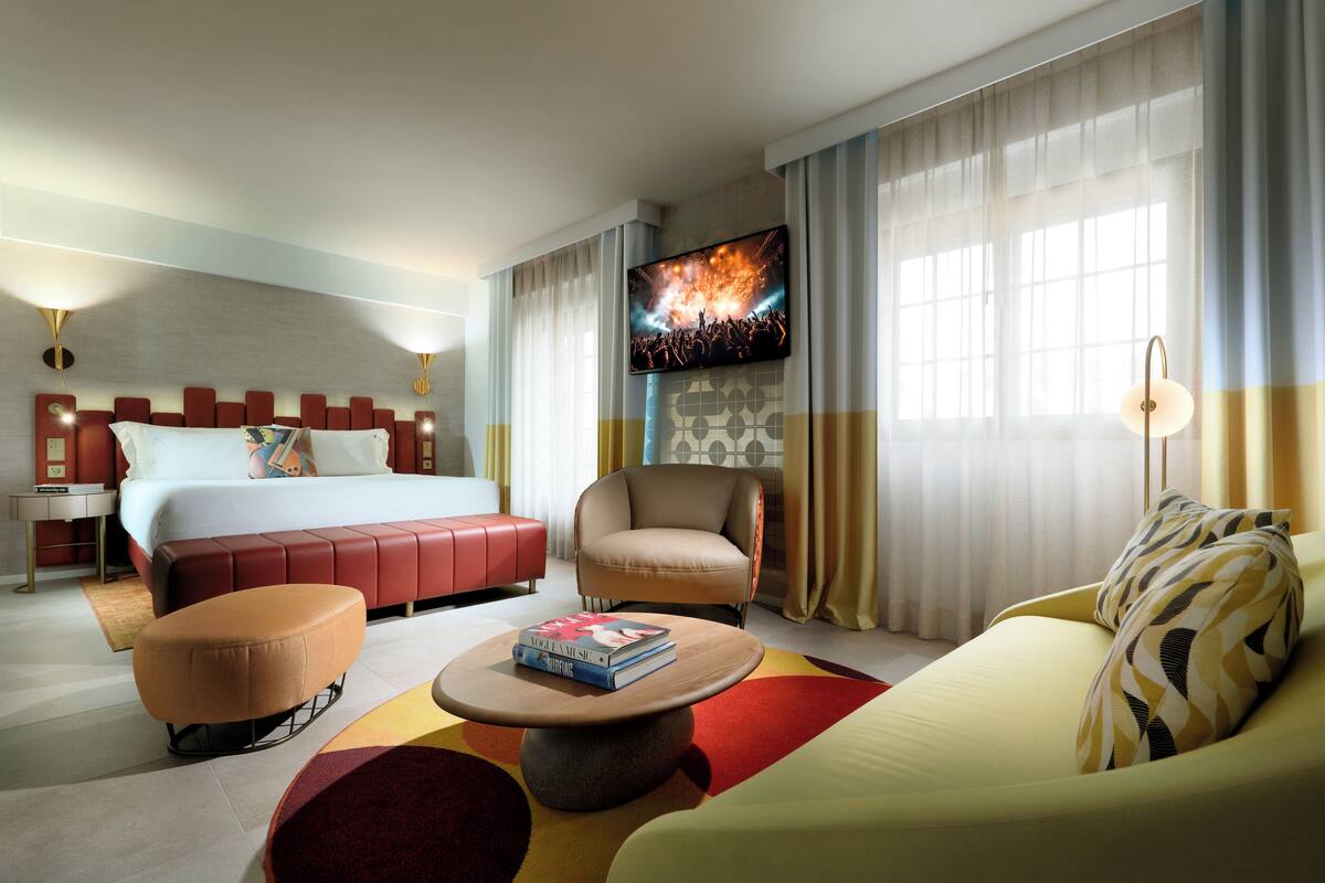Una habitación de hotel con una cama grande, un sofá, una silla, una mesita de centro y un televisor montado en la pared.