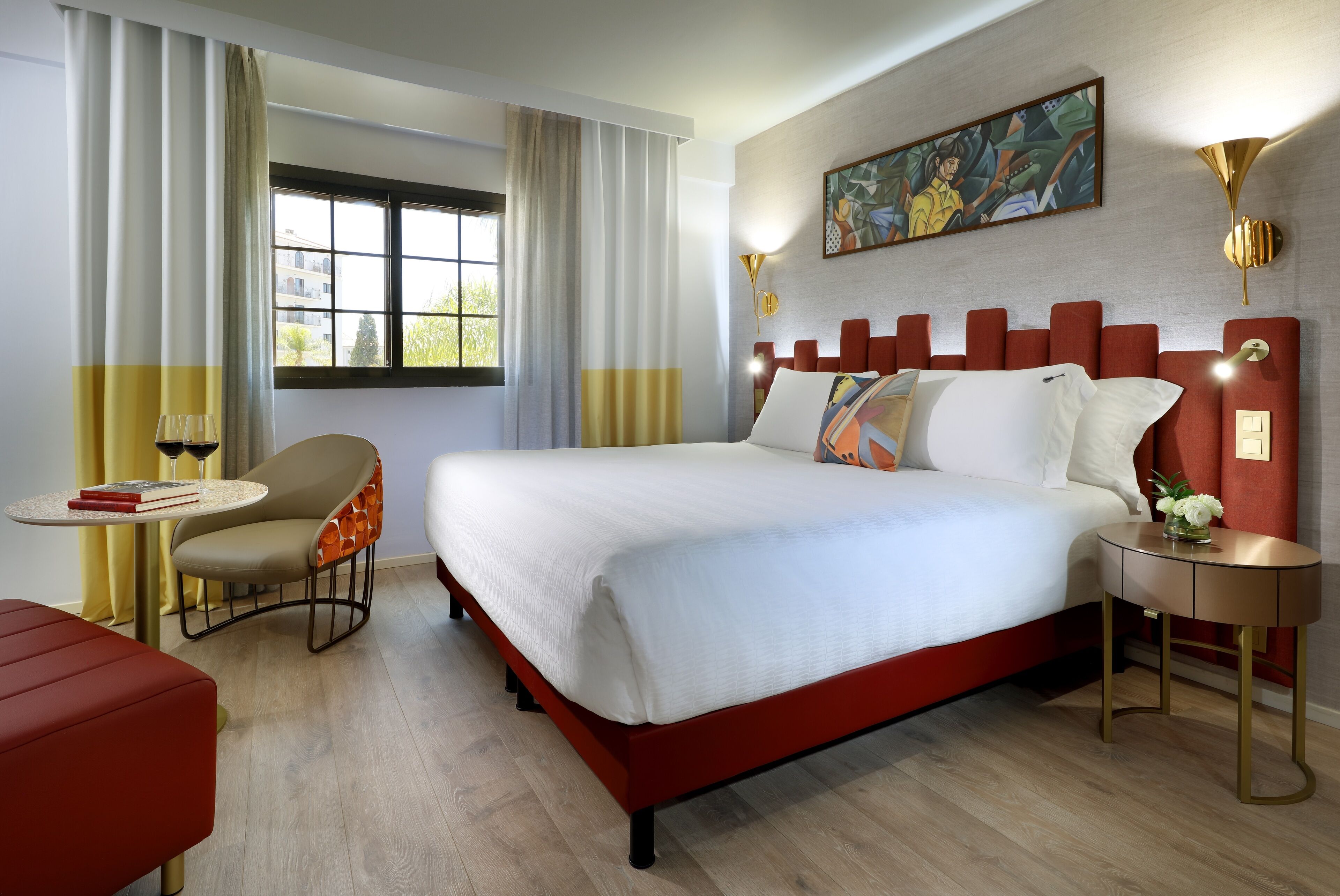 A modern hotel room with a large bed, a small table with a chair, a red sofa, and a painting on the wall.