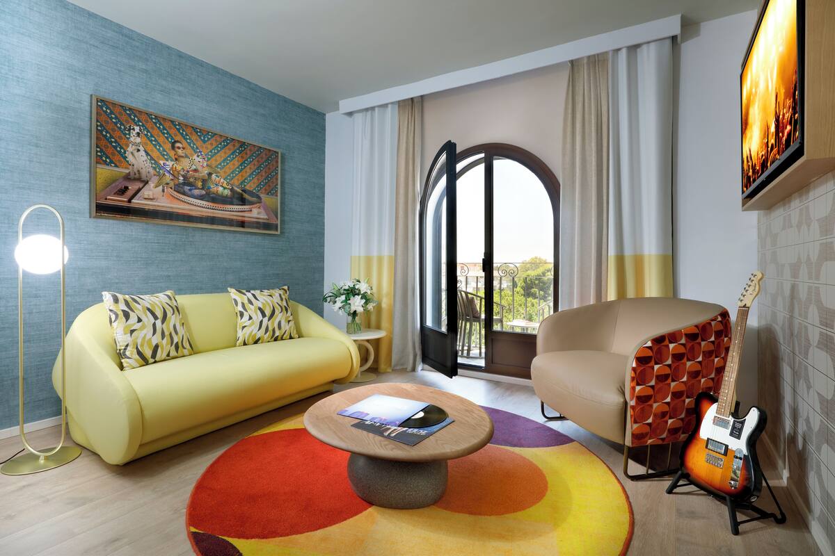 Una sala de estar moderna con un sofá amarillo, una mesa redonda de centro, una guitarra y un ventanal con cortinas.