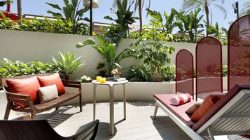 An outdoor patio with a wooden bench, a lounge chair, and a small table with a drink and flowers.