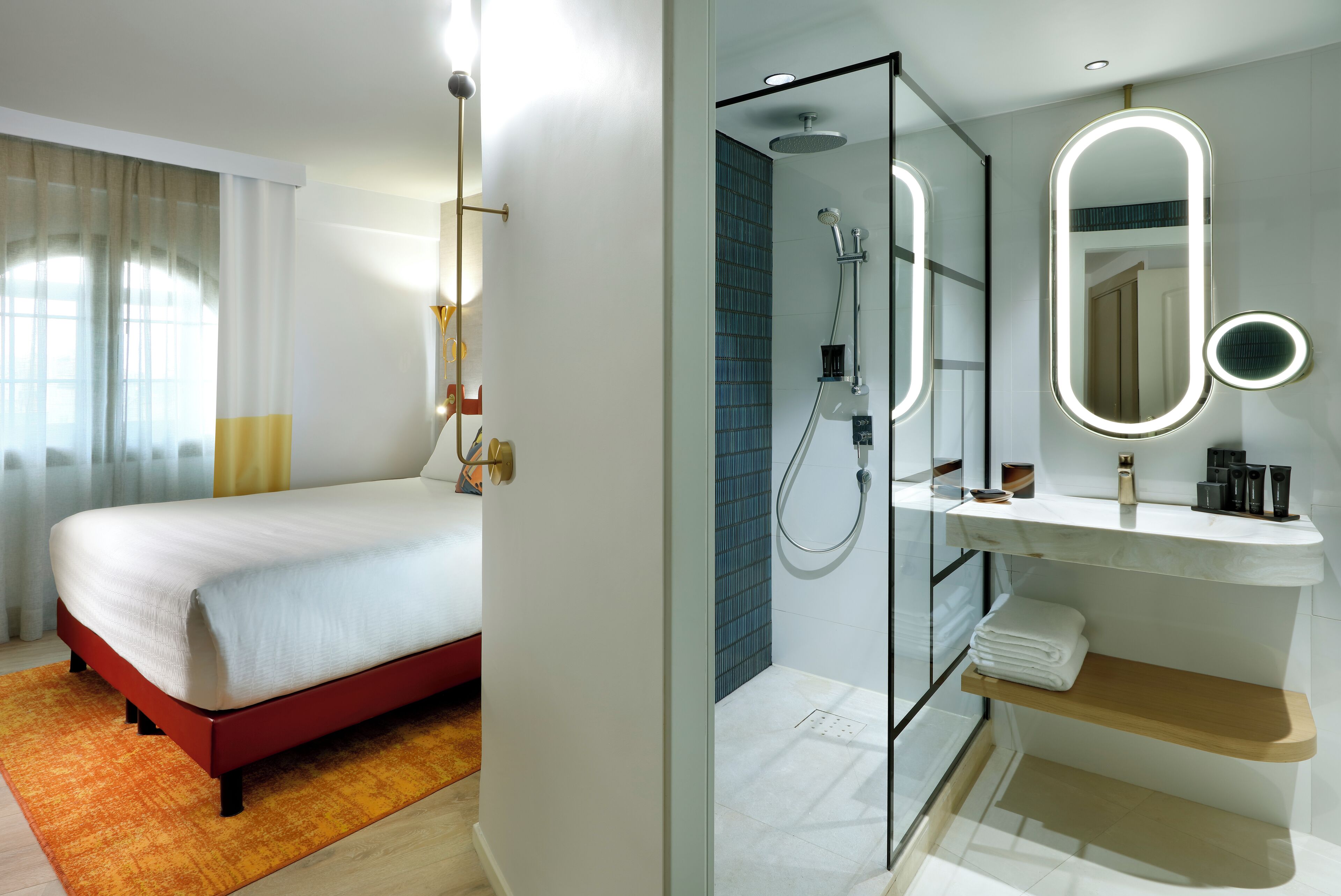 A hotel room with a bed, a bathroom with a shower, a mirror, and a shelf with toiletries.