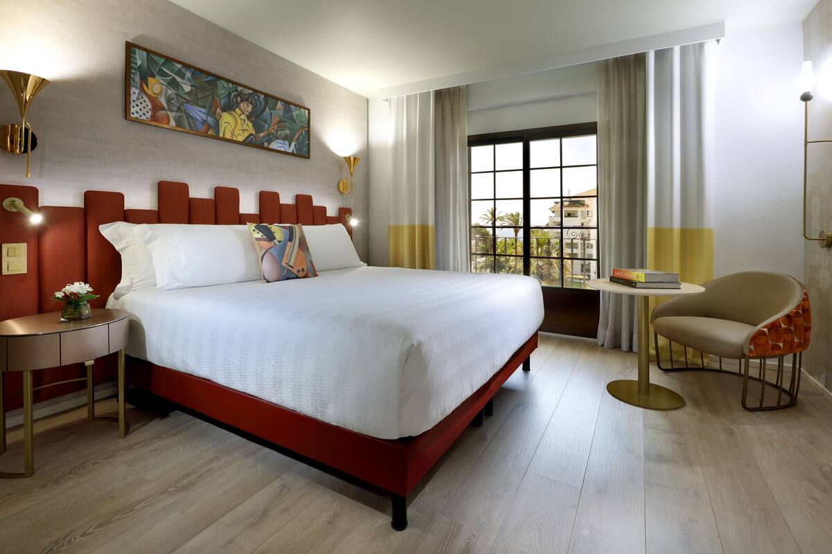 Una habitación de hotel moderna con una cama grande, una mesita de noche con lámpara, una silla y un ventanal con cortinas.