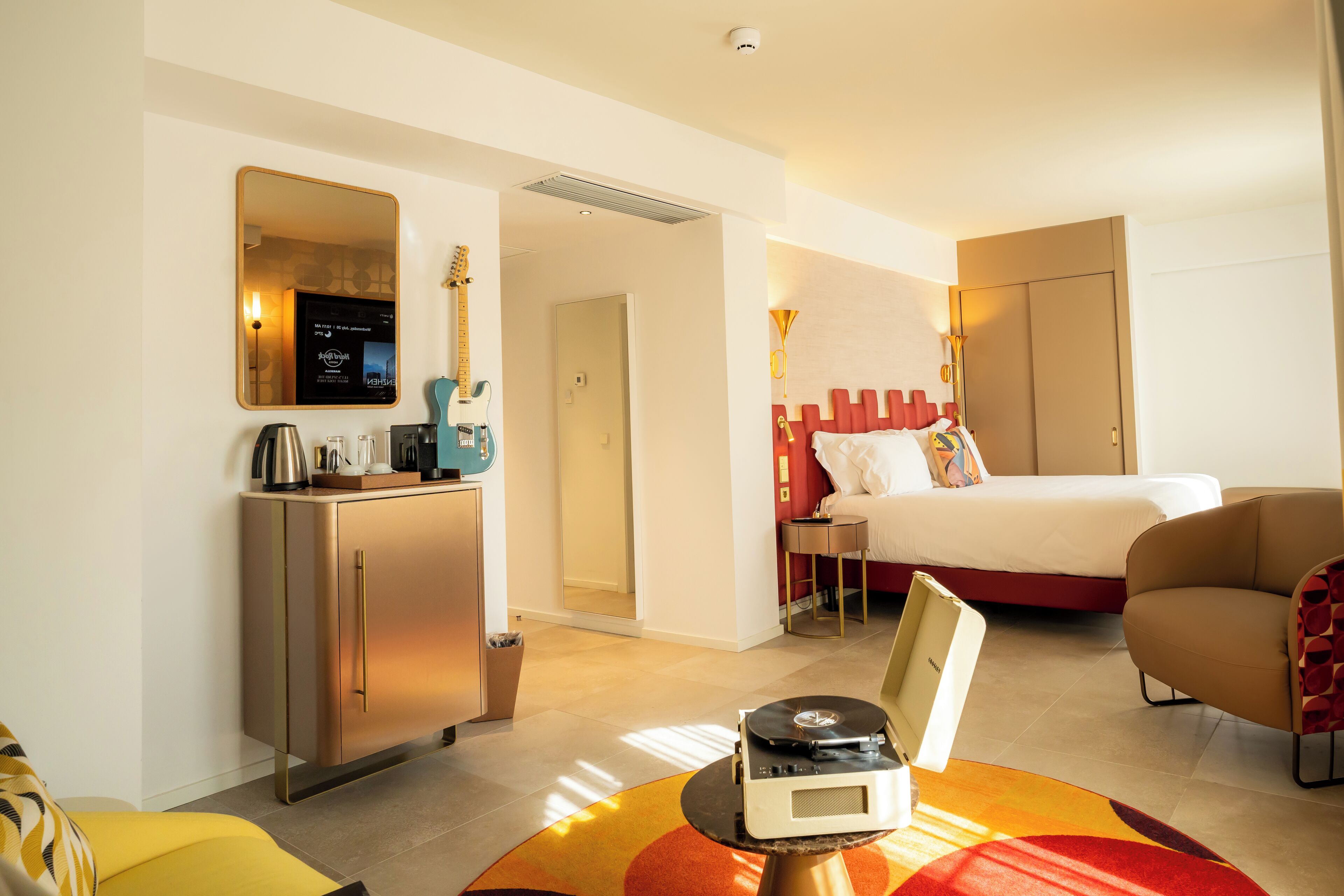 A hotel room with a bed, a desk with a computer, a chair, and a small table with a record player.