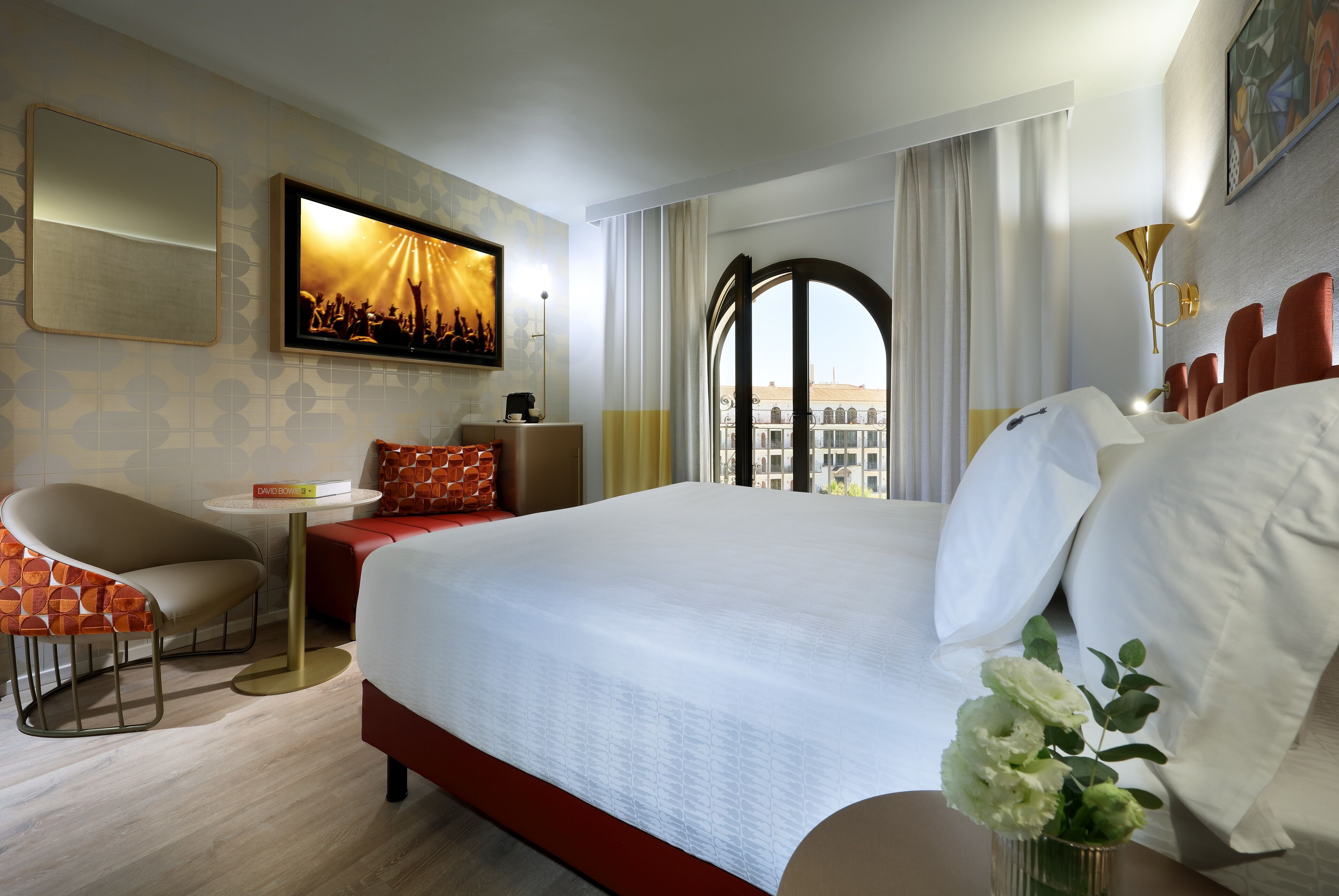 Hotelkamer met een groot bed, een aan de muur bevestigde televisie, een kleine tafel met stoel en uitzicht op gebouwen door een raam.