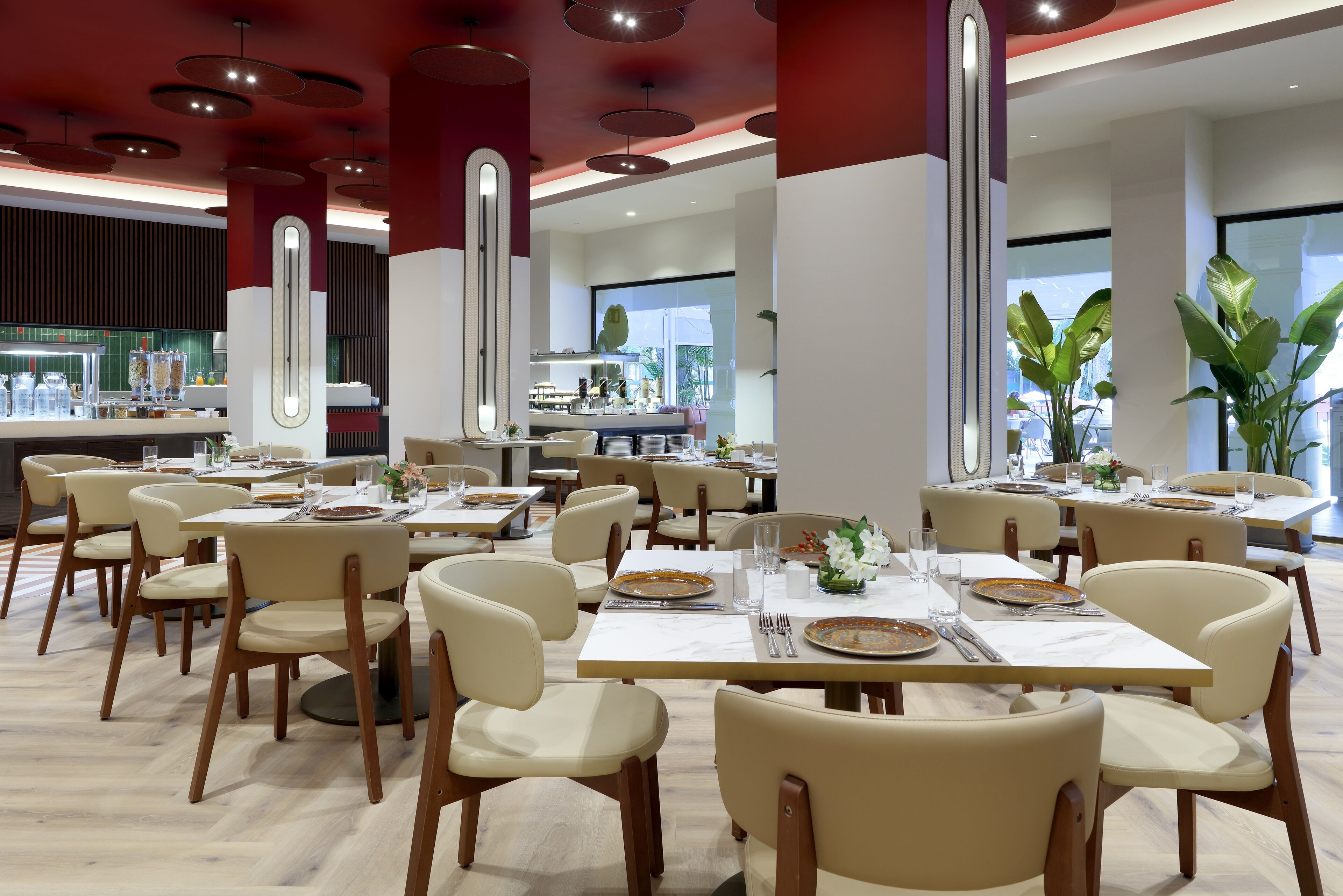A modern dining area with multiple tables and chairs, a kitchen in the background, and large windows allowing natural light.