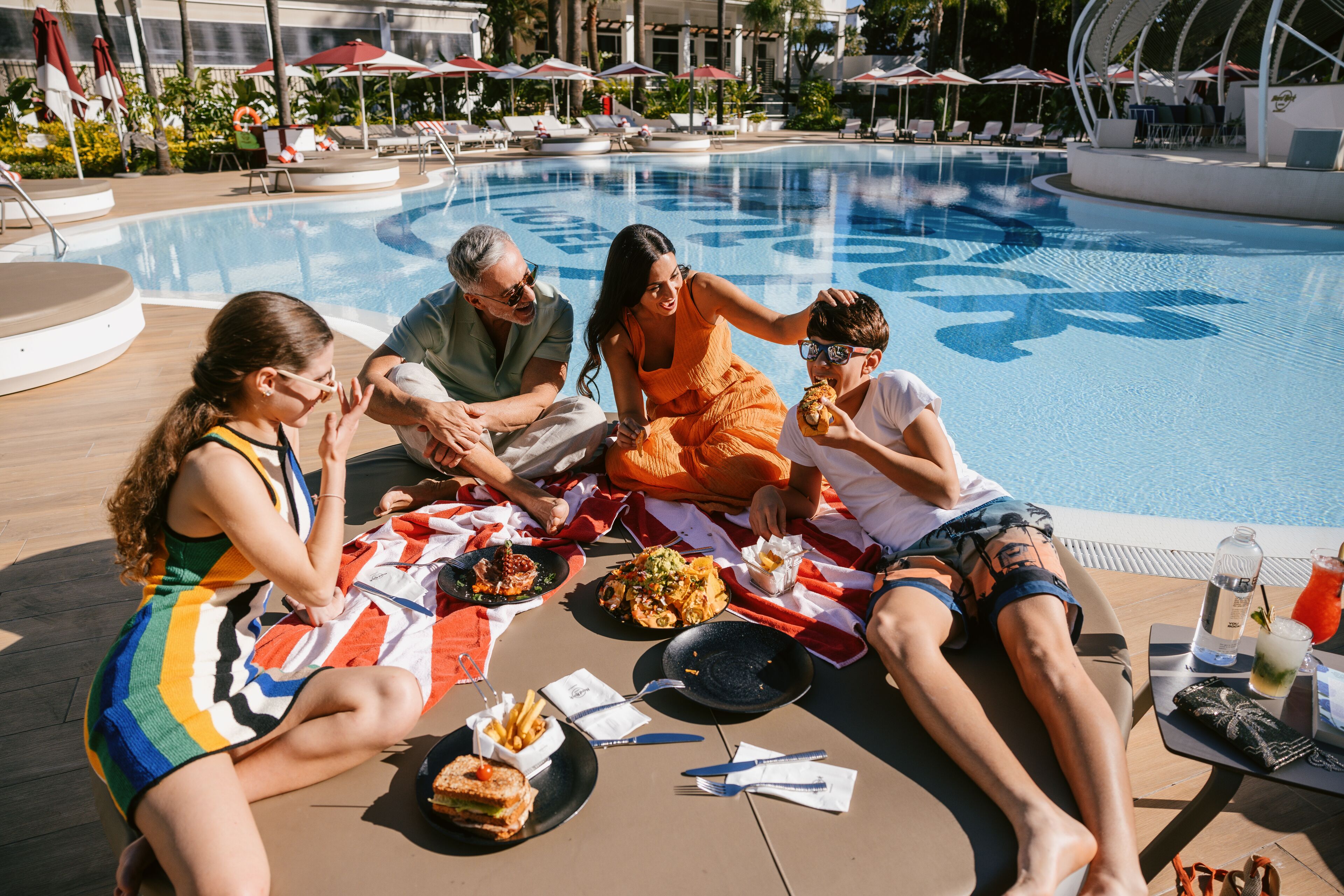 Un groupe de personnes est réuni autour d’une table pour dîner près d’une piscine, avec la piscine et des chaises longues en arrière-plan.