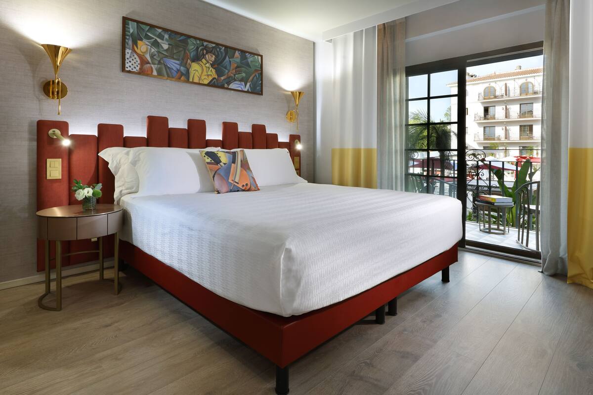 Una habitación de hotel moderna con una cama grande, una mesita de noche redonda y vistas al balcón exterior.