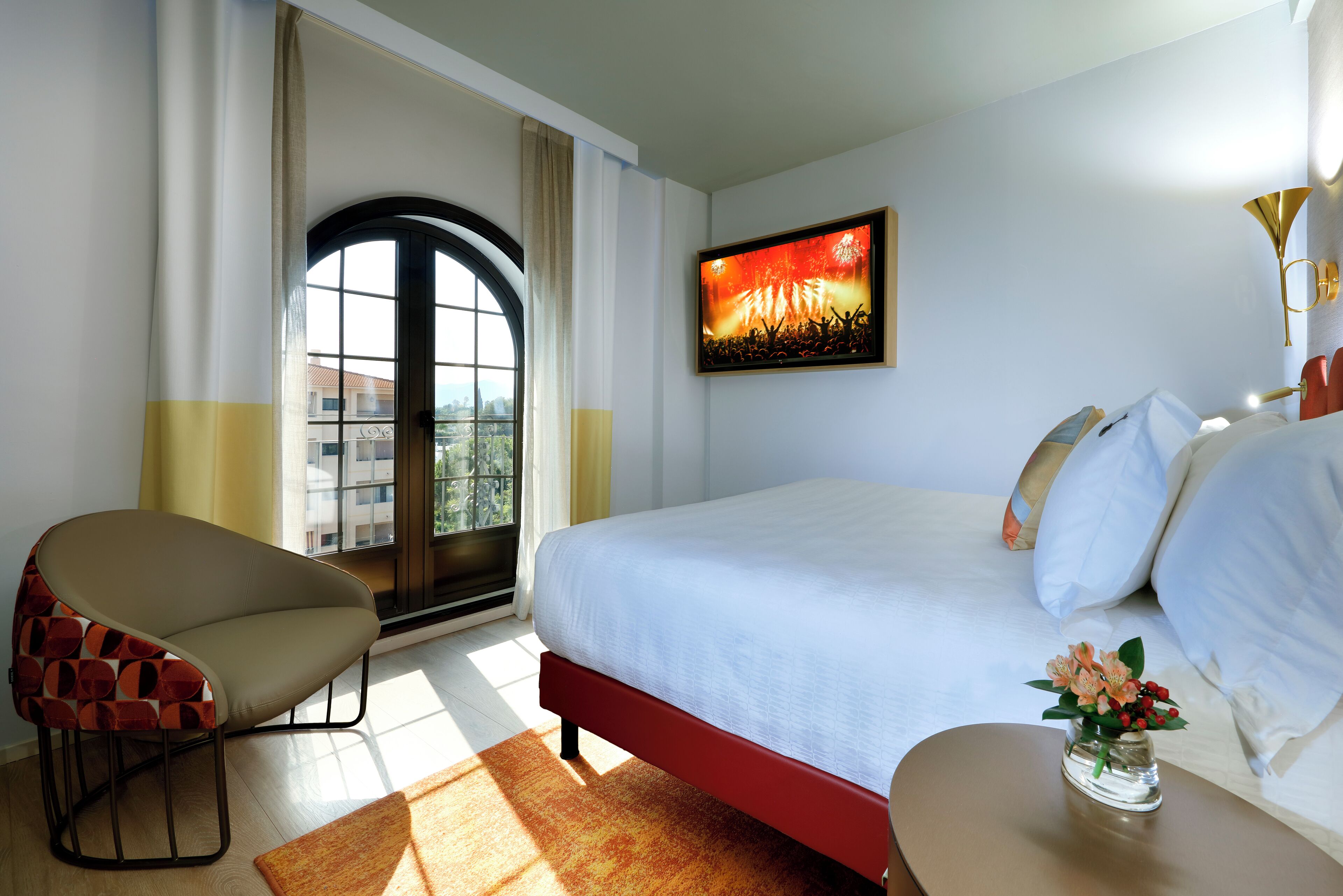 Habitación de hotel con cama, una silla, una mesita con flores, un cuadro en la pared y una ventana con cortinas.