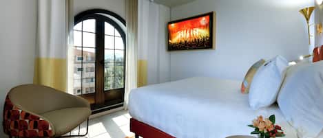 Ein Hotelzimmer mit Bett, Sessel, kleinem Tisch mit Blumen, einem Bild an der Wand und einem Fenster mit Vorhängen.