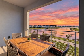A balcony with a wooden table and wicker chairs, overlooking a body of water and houses.