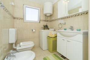 Een badkamer met toilet, wastafel en bad. Er is een raam, een handdoekhouder en een groene prullenbak.