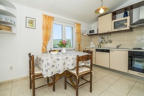 Cozinha compacta com área de refeições, apresentando uma mesa com toalha de mesa florida, cadeiras de madeira, uma janela com cortinas e eletrodomésticos básicos.