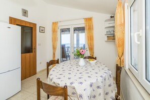 Uma sala de jantar com uma mesa redonda, cadeiras, um vaso com flores e vista para o mar através de uma porta de vidro.