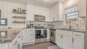 A modern kitchen with white cabinetry, a stainless steel microwave, a built-in oven, and a dishwasher. There are wooden shelves above the counter and a window with a view of palm trees.