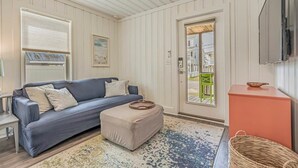 A room with a dark blue sofa, a beige ottoman, a patterned rug, a wooden floor, a white door, and a television mounted on the wall.