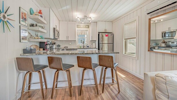 A modern kitchen with white cabinetry, a central island with bar stools, and a view of the beach through a window. - Coconut Flats Duplex- Upstairs- (North Myrtle Beach)