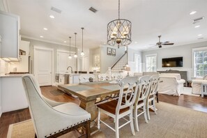 A modern kitchen with white cabinets, a dining area with a wooden table and white chairs, a staircase, and a living room with a TV and seating area. - 🌴 Moon Landing • Luxury WaterSound West Retreat with LSV, Bikes & Pool Access (30A WaterSound)