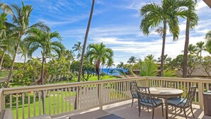 A balcony with a table and chairs, overlooking a lawn with palm trees and a view of the ocean.